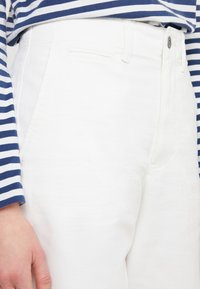 Närbild av en person som bär vita högmidjade byxor med knapp och en marinblå och vit randig långärmad tröja synlig högst upp.