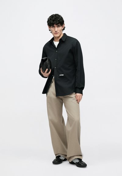 Ung mand med krøllet hår iført sort skjorte med knapper, beige bukser med vide ben, stribede sneakers, holder en sort tekstureret clutchtaske.