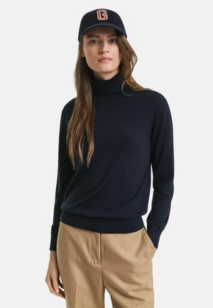 Marineblauer Rollkragenpullover mit langen Ärmeln, glatter Textur und eng anliegenden Bündchen, kombiniert mit beigen Hosen und einer marineblauen Mütze mit einem orangefarbenen Logo.