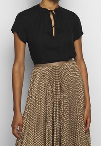 Mustamusta blouse, jossa on avaimenrei'ityyppinen yksityiskohta ja nauhat kaula-aukossa, yhdistettynä ruskeaan houndstooth-kuvioituun laskostettuun hameeseen, jossa on satiinivyötärö.