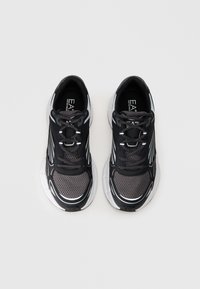 Sapatilhas de desporto em preto e cinza com parte superior em malha, bico arredondado e sola branca espessa. Apresentam atacadores e detalhes em branco contrastantes.