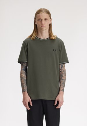 Jeune homme aux longs cheveux blonds et tatouages couvrant tout le bras, portant un T-shirt vert olive et un pantalon noir, debout devant un fond uni.