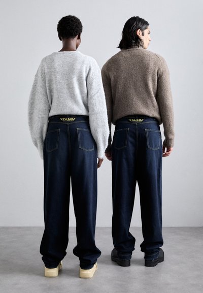 Two people, one in a gray fluffy sweater and the other in a brown sweater, both wear dark blue, loose-fitting jeans with yellow branding on the waistband.