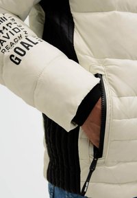 Person wearing a beige and black quilted jacket with zipper pocket, hand inside pocket, and text "GOAL" on the sleeve.
