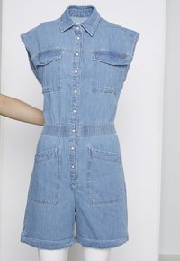 Combinaison en denim bleu clair, dotée d'un col, de manches courtes retroussées, d'une fermeture boutonnée à l'avant et de deux poches poitrine. Taille élastique.