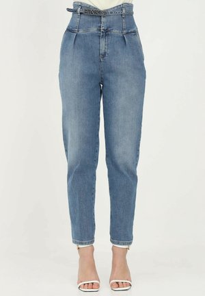 Vaqueros azules de talle alto y corte estrecho con pliegues y un cinturón de marca, combinados con sandalias blancas de tacón alto y puntera abierta.