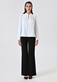 Blusa bianca con scollatura a chiave e maniche lunghe, abbinata a pantaloni neri a gamba larga e scarpe nere con punta.