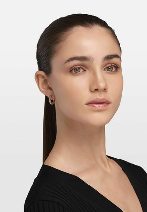 Goldene Creolen mit strukturiertem Design, am linken Ohr getragen. Glatte, helle Haut mit minimalem Makeup; dunkles, glattes Haar nach hinten gebunden.