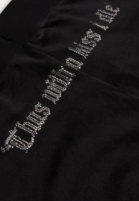 Crni tekstilni odjevni predmet s teksturiranom površinom i sjajnim ukrasima od rhinestona koji čine zakrivljeni dizajn teksta.