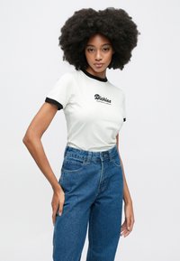 Λευκό μπλουζάκι με μαύρο ρέλι στο λαιμό και κοντά μανίκια, με μαύρη επιγραφή "Dickies Workwear Company" στο στήθος, σε συνδυασμό με μπλε ψηλόμεσα τζιν.