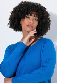 Blaues Langarmoberteil aus glattem Stoff, mit rundem Ausschnitt und leichtem Struktur. Das Model posiert mit der Hand am Kinn vor einem weißen Hintergrund.