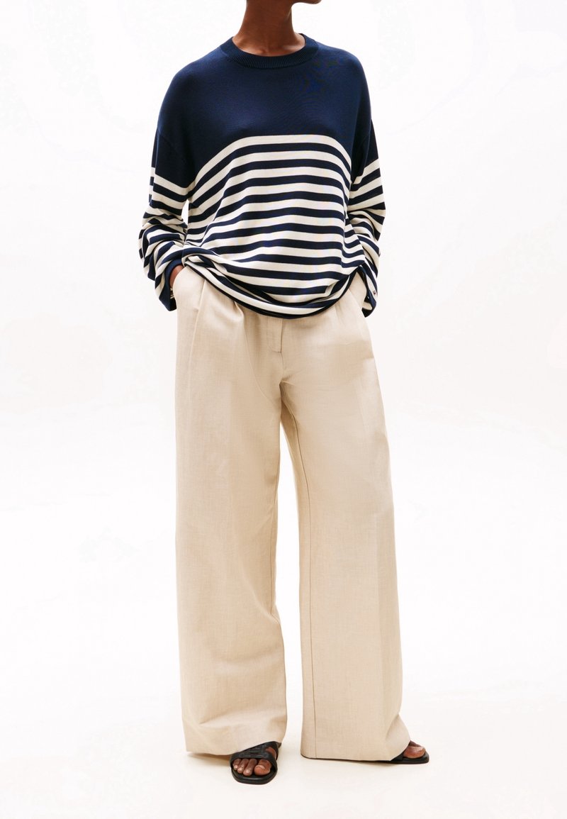 Personne portant un pull rayé bleu marine et blanc, un pantalon beige à jambes larges, des sandales noires ouvertes aux orteils, debout les mains dans les poches.