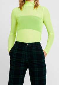 Personne portant un haut long à manches transparentes vert fluo sur un bandeau assorti, associé à un pantalon écossais noir et vert foncé, les mains dans les poches.