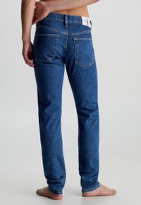 Barfota person som bär blå denimjeans med synliga bakfickor och en vit Calvin Klein-logotypapplikation vid midjan.