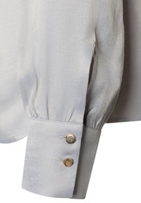 Hellgraue Bluse mit seidiger Textur, die einen gerundeten Ärmelabschluss und zwei goldene Knöpfe zum Verschließen aufweist. Dezenter Glanz und geraffte Stoffdetails.