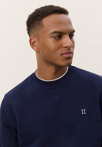 Temno modra majica z dolgimi rokavi iz bombažnega materiala, z okroglo izrezanim vratom, rebrastimi manšetami in majhno belo vezeno podrobnostjo "II" na prsih.