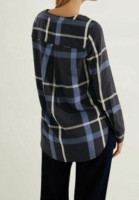 Femme portant une chemise ample à manches longues avec un large motif à carreaux bleu marine, bleu clair et blanc, debout avec le dos tourné vers le spectateur.
