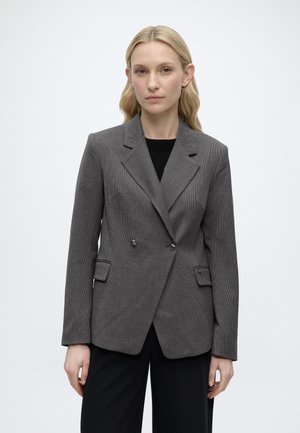 Femme blonde portant un blazer gris à carreaux à double boutonnage et un pantalon noir, debout devant un fond clair uni.