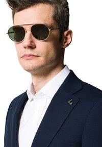 Okulary przeciwsłoneczne w złotej oprawie z zielonymi szkłami noszone przez mężczyznę w granatowym żakiecie. Żakiet ma subtelny logo na klapie i białą koszulę pod nim.