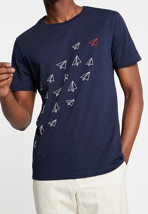 Mann trägt ein marineblaues T-Shirt mit weißem und einem roten Papierflugzeug-Grafik, die diagonal von unten links nach oben rechts fliegt.
