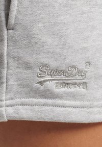 Telón gris que presenta un primer plano de un logo de SuperDry, bordado en un tono gris ligeramente más oscuro, detallando las costuras y la textura del material.