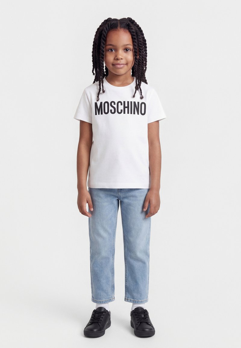 Νεαρή κοπέλα με πλεξούδες, φορώντας ένα λευκό t-shirt Moschino, ανοιχτό μπλε τζιν και μαύρα αθλητικά παπούτσια, στέκεται μπροστά σε λευκό φόντο.