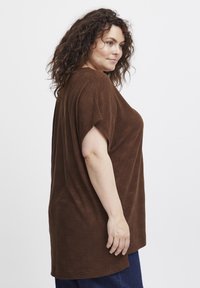 Brun tekstureret oversized top med korte ærmer og rund halsudskæring, parret med mørkeblå jeans. Har subtile bølgemønstre.