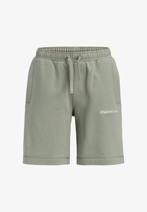 Garçon aux cheveux bouclés portant un t-shirt et un short assortis vert sauge, des chaussettes blanches et des baskets blanches, debout devant un fond uni.