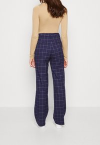 Kvinna i marinblå vida byxor med beige rutmönster och en figursydd långärmad beigetopp, stående mot en enkel vit bakgrund.