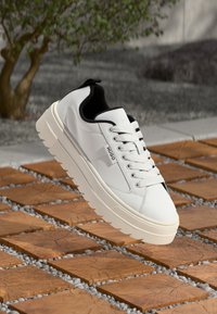 Biała sneakersy z gładką skórzaną cholewką, grubą gumową podeszwą z zigzagowym wzorem, czarnym wnętrzem i subtelnym logo po boku.