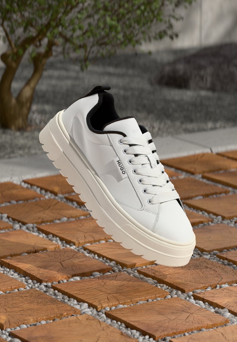 Biała sneakersy z gładką skórzaną cholewką, grubą gumową podeszwą z zigzagowym wzorem, czarnym wnętrzem i subtelnym logo po boku.