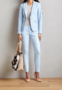 Blazer bleu clair sur mesure et pantalon assorti, complétés par un haut beige. Écharpe brune et blanche à motif nouée sur un sac fourre-tout beige. Talons roses.