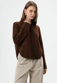 Brauner Strickpullover mit rundem Ausschnitt, geripptem Saum und seitlicher Knopfverzierung. Kombiniert mit beigen Hosen, präsentiert vor schlichtem Hintergrund.