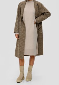 Cappotto lungo a quadri marroni sopra un vestito a coste beige, abbinato a stivaletti alla caviglia con suola chunky beige chiaro.