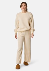 Vrouw die staat in een crèmekleurige, geribbelde top met lange mouwen en bijpassende broek, met bruine instapslippers op een witte achtergrond.
