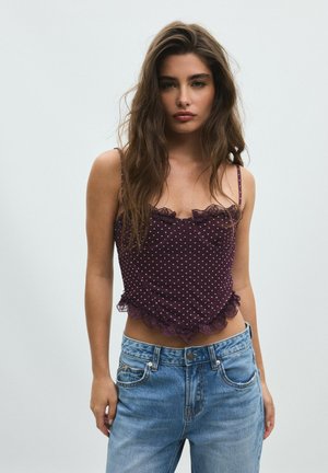 Jeune femme aux longs cheveux bruns portant un caraco violet à pois orné de dentelle et un jean bleu, debout devant un fond clair uni.