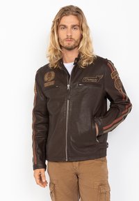 Giacca da moto in pelle marrone con zip laterali, dettagli in patch e strisce rosse e marroni lungo le maniche. Texture liscia, colletto alla mandarino.