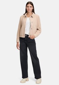 Kobieta stojąca w beżowej zapinanej marynarce, białej bluzce, ciemnych szerokich dżinsach i jasnych botkach na obcasie, na jednolitym tle.