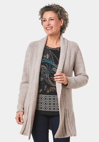 Hellbeige langer Cardigan mit offenem Vorderteil, geripptem Textur und Schalkragen, kombiniert mit einem gemusterten schwarzen Top und dunklen Jeans.