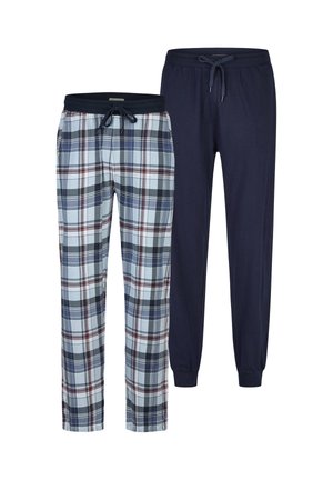 Man zonder shirt met tribal tatoeage op de linkerarm, gekleed in blauw-witte geruite slaapbroek, staat op blote voeten en kijkt opzij met een lichte glimlach.