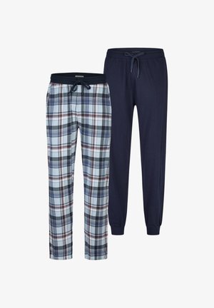 Oberkörperfreier Mann mit tribalem Tattoo am linken Arm, trägt blaue und weiße karierten Schlafanzughosen, steht barfuß und schaut mit einem leichten Lächeln zur Seite.