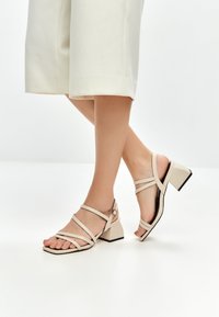 Sandales beige à lanières avec un talon épais, dotées d'un bout carré, d'une texture en cuir lisse et d'une bride de cheville réglable.