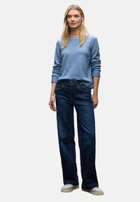 Hellblauer strukturierter Strickpullover kombiniert mit dunklen, weit geschnittenen Jeans und beigen Schuhen. Das Outfit zeichnet sich durch einfache, klare Linien aus.