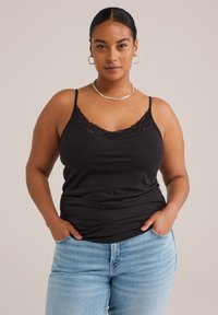Frau in schwarzem, mit Spitze verziertem Top, hellblauen Jeans, silberner Halskette und Creolen, steht mit Händen in den Taschen vor einem schlichten Hintergrund.