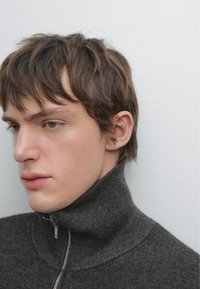Mörkgrå zip-sweater med hög krage; ribbad textur; personen som bär den har kort, rufsig hår och ljus hudton.
