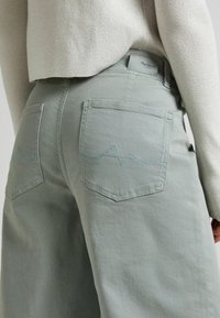 Vaaleanvihreät farkut korkealla vyötäröllä, joissa on kaksi takataskua, joissa on sinisellä langalla tehty aalto-kuvio. Kangas vaikuttaa sileältä ja hieman jämäkältä.