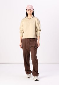 Felpa beige con zip a quarto e polsini a costine, abbinata a pantaloni cargo marroni a vestibilità ampia e a un cappellino da baseball rosa chiaro.