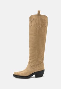 Bota vaquera de ante color beige hasta la rodilla, con costuras decorativas, punta afilada y un tacón bloque moderado. Presenta una pestaña para tirar en la parte superior.