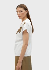 T-shirt bianco in cotone con una vestibilità rilassata, caratterizzato da un design animale con perline dorate sulla spalla. Maniche corte e scollo rotondo.