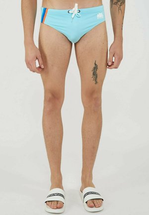 Uomo che indossa slip da bagno azzurri chiari con strisce arancioni e blu, coulisse bianca, tatuaggio nero sulla coscia sinistra e ciabatte bianche Emporio Armani.